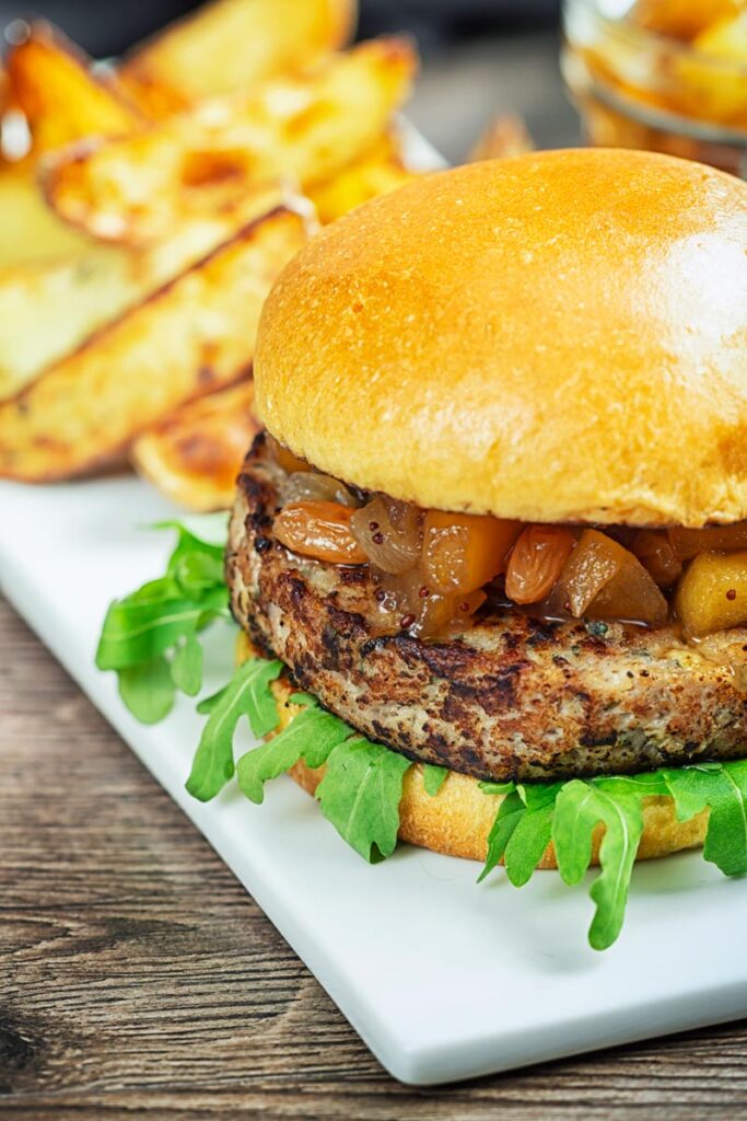 Portrait image of a pork and apple burger served on a white rectangle plate with a chutney topping and potato wedges