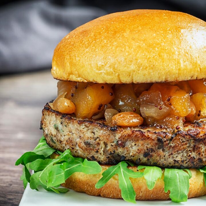 Square image of a pork and apple burger served on a white plate with a chutney topping