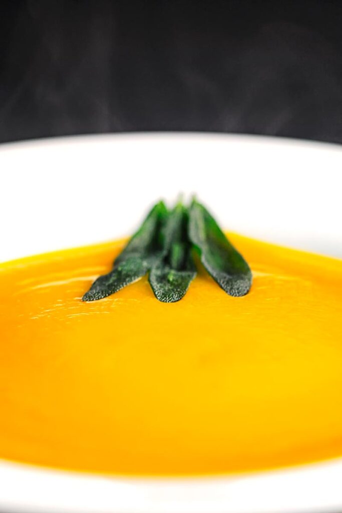 Portrait close image of a bowl of silky steaming hot roasted butternut squash soup served with fried sage leaves