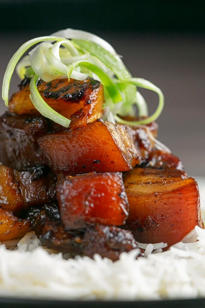 Portrait close up image of a single serving of glazed sticky pork belly with pineapple on a bed of rice and garnish of shredded green onion