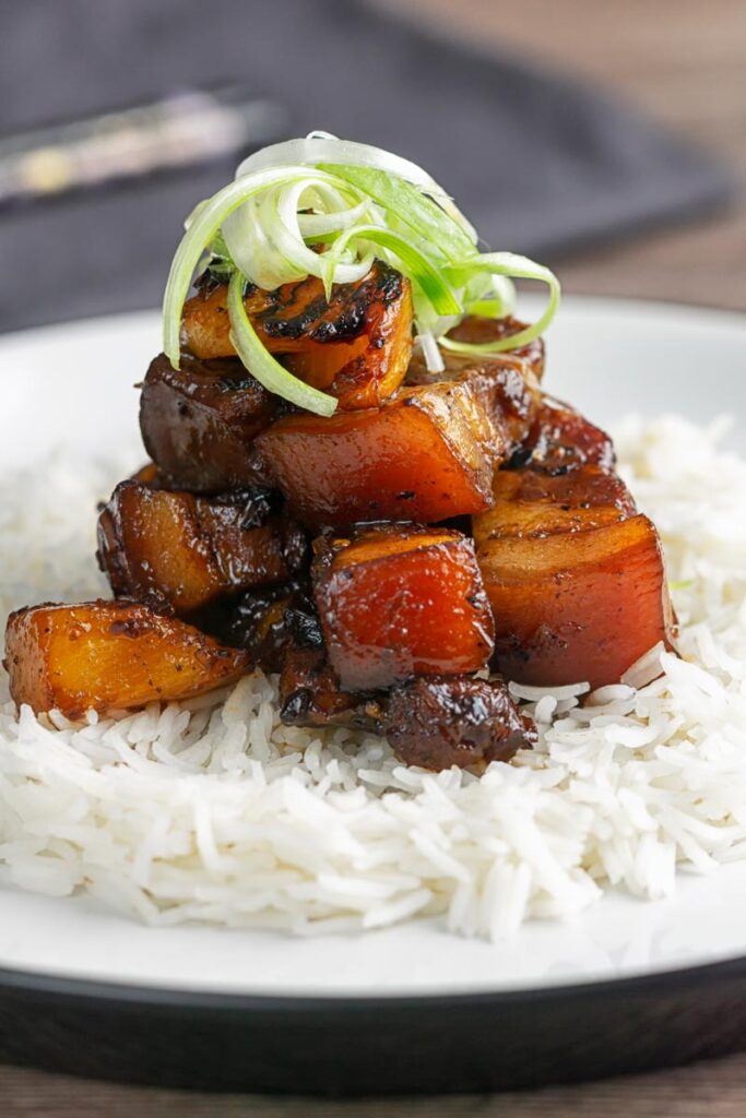 Portrait image of a single serving of glazed sticky pork belly with pineapple on a bed of rice and garnish of shredded green onion on a white plate