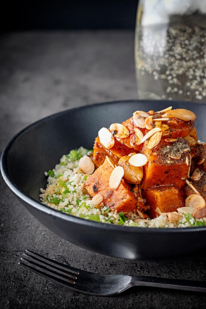 Portrait image of a vegetarian sweet potato tagine garnished with flaked almonds and zaatar on a bed of couscous