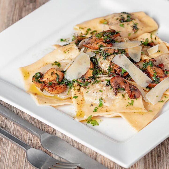 Square image of mushroom stuffed ravioli with balsamic mushrooms and Parmesan cheese shavings served on a white square plate