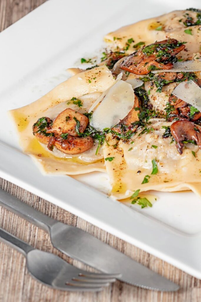 Portrait image of mushroom stuffed ravioli with balsamic mushrooms and Parmesan cheese shavings served on a white square plate