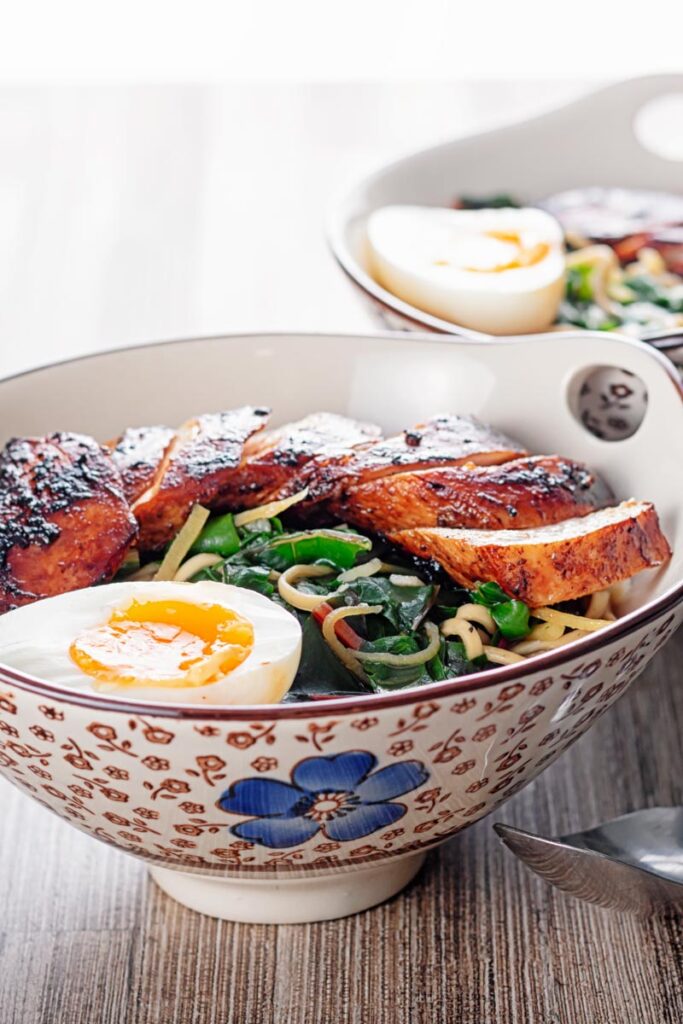 Square image of a hoisin chicken ramen with a boiled egg and Swiss chard served in an Asian style bowl decorated with a blue flower