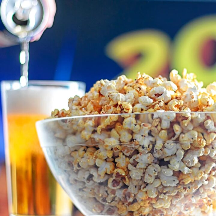 Square image of spiced popcorn in a glass bowl with a beer being poured in the background