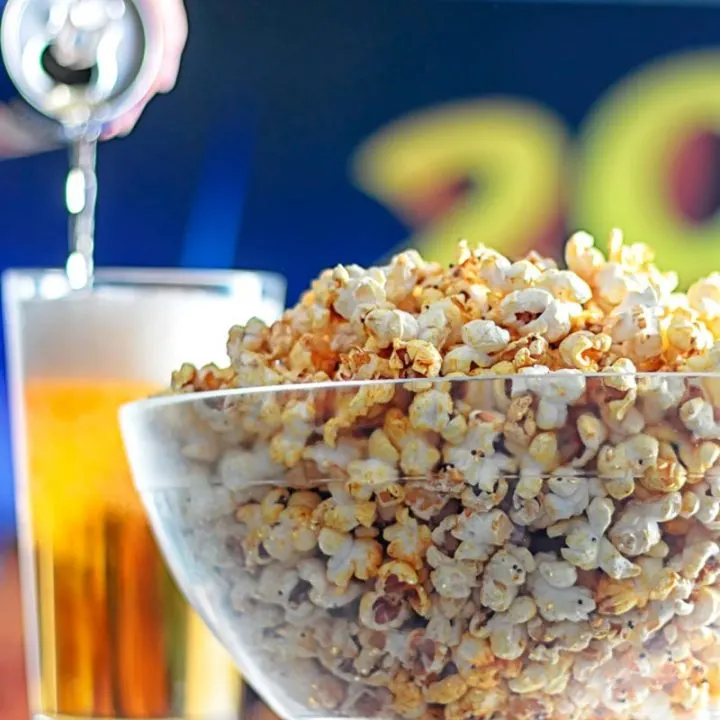Square image of spiced popcorn in a glass bowl with a beer being poured in the background