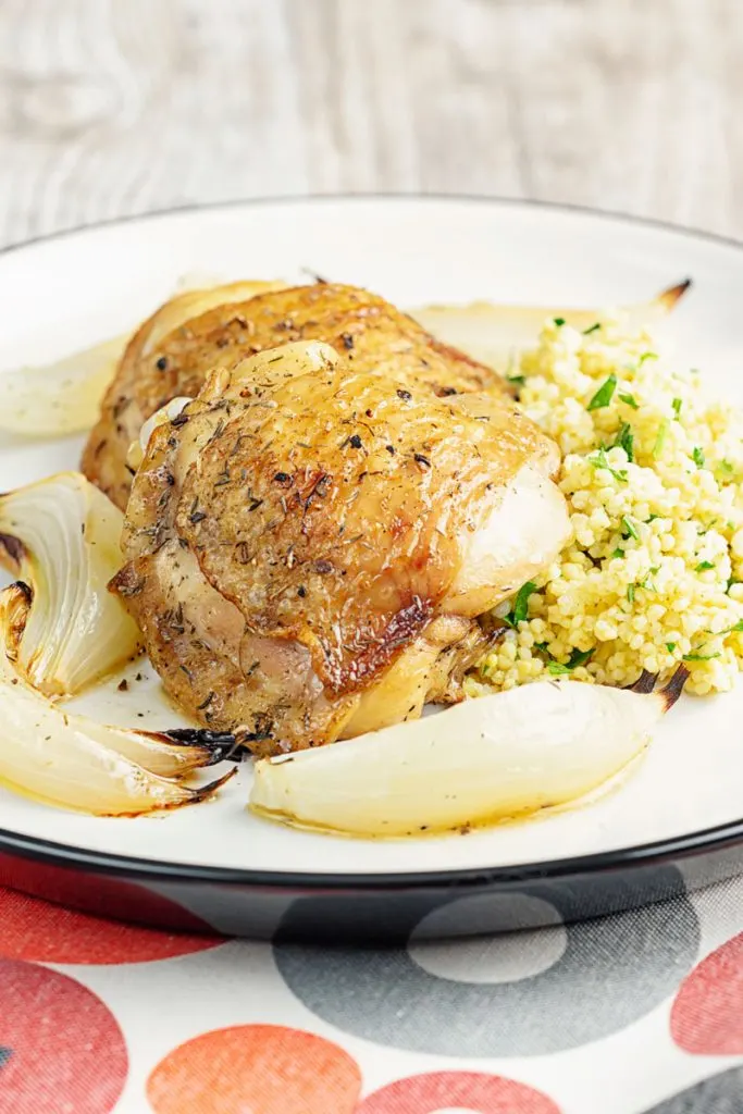 Portrait close up image of lemon and garlic chicken thighs served with roasted onions and a millet side dish served on a white plate