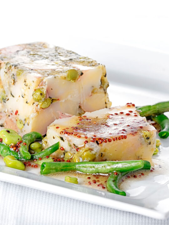 Portrait image of a vegetable terrine featuring peas and potatoes served on a white plate with beans and a mustard dressing