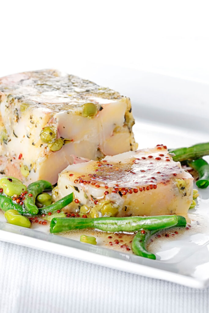 Portrait image of a vegetable terrine featuring peas and potatoes served on a white plate with beans and a mustard dressing