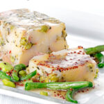 Portrait image of a vegetable terrine featuring peas and potatoes served on a white plate with beans and a mustard dressing with a text overlay