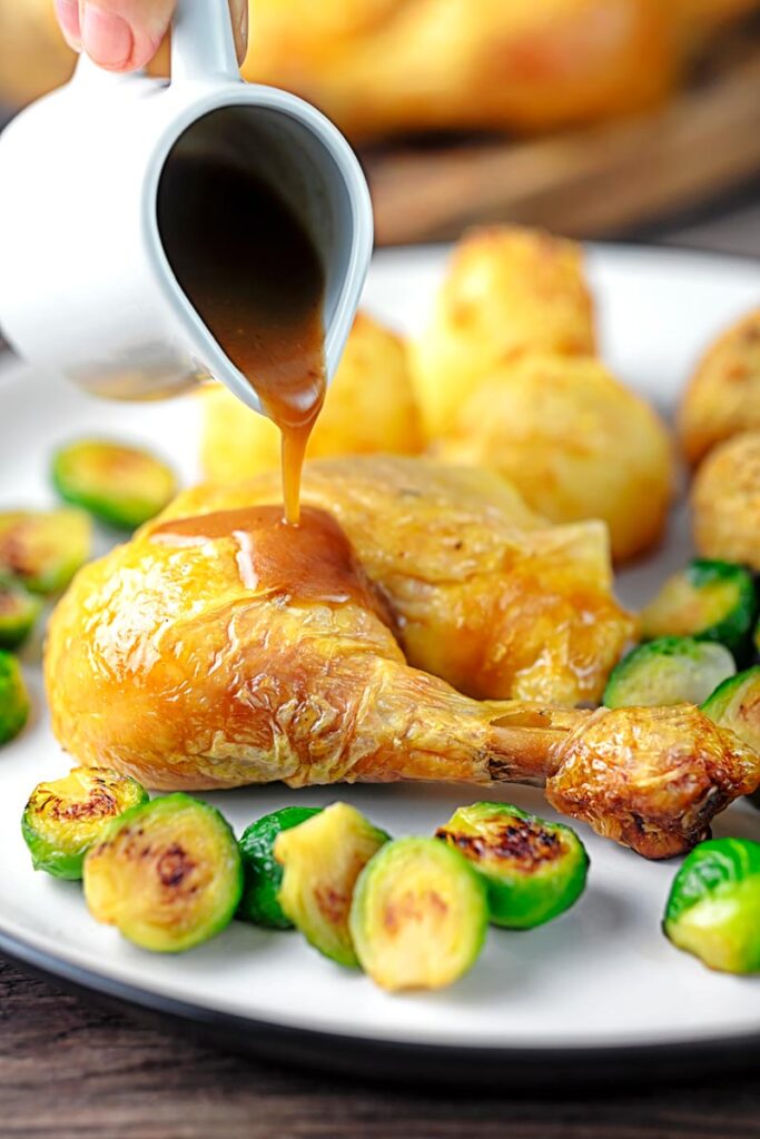 A perfect silky pan gravy being poured of over a roast chicken leg served with Brussels sprouts and roast potatoes
