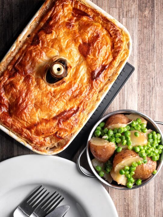 Portrait overhead image of a pork and apple pie with a puff pastry crust served with buttered peas and potatoes