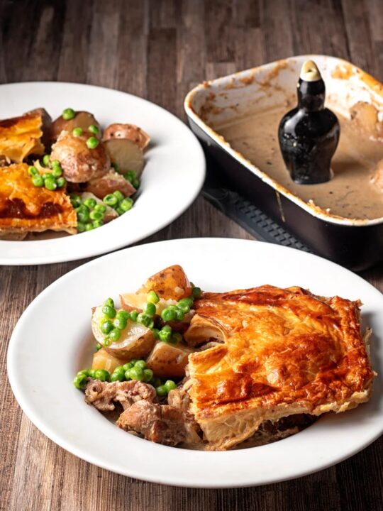 Portrait image of a pork and apple pie served in a white bowl with peas and potatoes and and empty pie dish