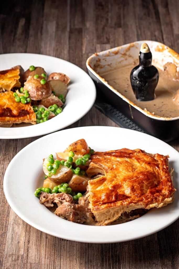 Portrait image of a pork and apple pie served in a white bowl with peas and potatoes and and empty pie dish