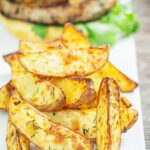 Portrait image of potato wedges served on a white plate with a burger in the background