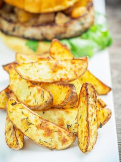 Portrait image of potato wedges served on a white plate with a burger in the background