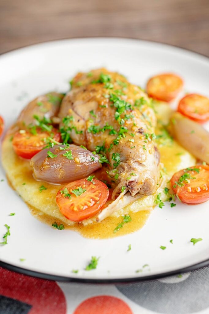 Portrait image of rabbit cacciatore featuring rabbit legs, shallots and cherry tomatoes served on cheesy polenta with lots of parsley
