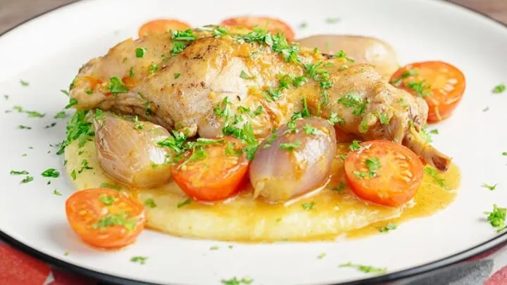 Landscape image of rabbit cacciatore featuring, rabbit legs shallots and cherry tomatoes served on cheesy polenta with lots of parsley