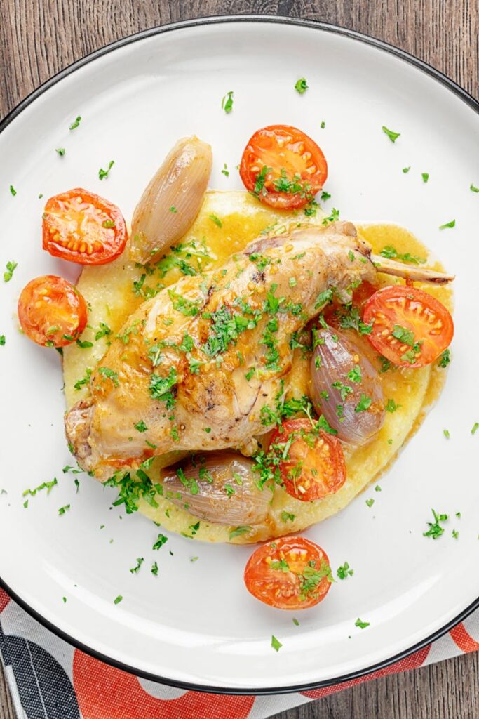 Square overhead image of rabbit cacciatore featuring, rabbit legs shallots and cherry tomatoes served on cheesy polenta with lots of parsley