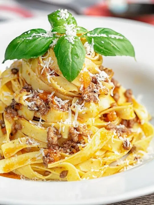 Portrait image of pasta served with a ragu bolognese meat sauce served in a white bowl with basil and grated Parmesan cheese