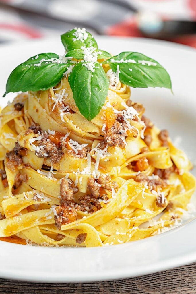Portrait image of pasta served with a ragu bolognese meat sauce served in a white bowl with basil and grated Parmesan cheese