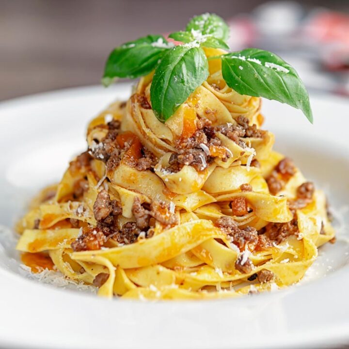 Square image of pasta served with a ragu bolognese meat sauce served in a white bowl with basil and grated Parmesan cheese