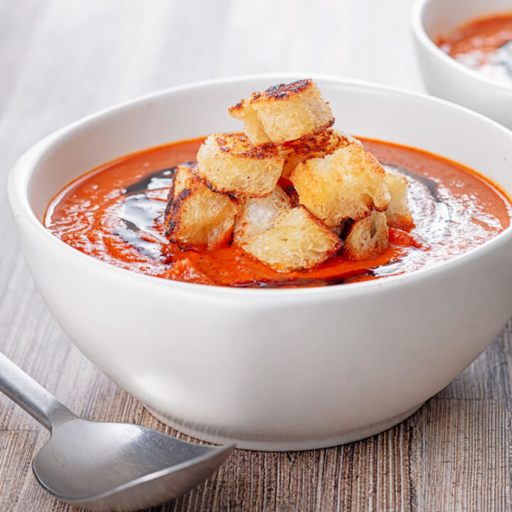 Square image of a bowl of roasted tomato soup served with a tower of toasted croutons and a balsamic swirl
