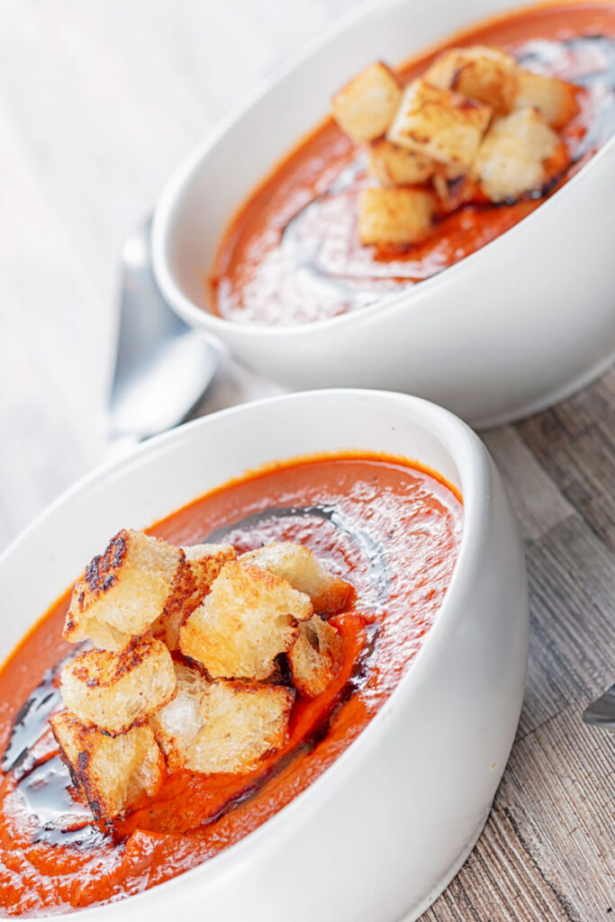 Portrait image of two bowls of roast tomato soup served with toasted croutons and balsamic swirl