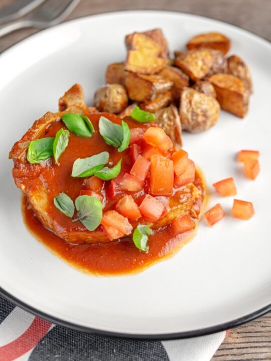 A portrait image of a slow cooker pork chop served on a white plate in a tomato sauce with tomato concasse and basil served with fried potatoes