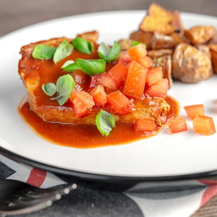Square image of a slow cooker pork chop served on a white plate in a tomato sauce with tomato concasse and basil served with fried potatoes with text overlay