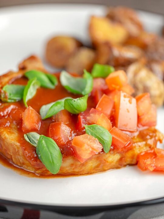 Portrait image of a slow cooker pork chop served on a white plate in a tomato sauce with tomato concasse and basil served with fried potatoes