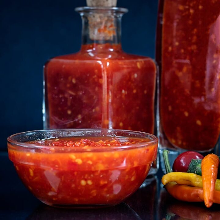Square image of sweet chili sauce served in a dippiing bowl with chillies and bottles of sauce