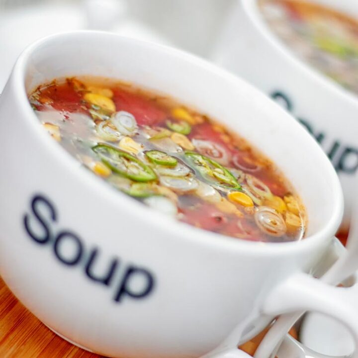 Square image of a tomato and sweetcorn broth based soup served in a white porcelain cup with soup printed on the front