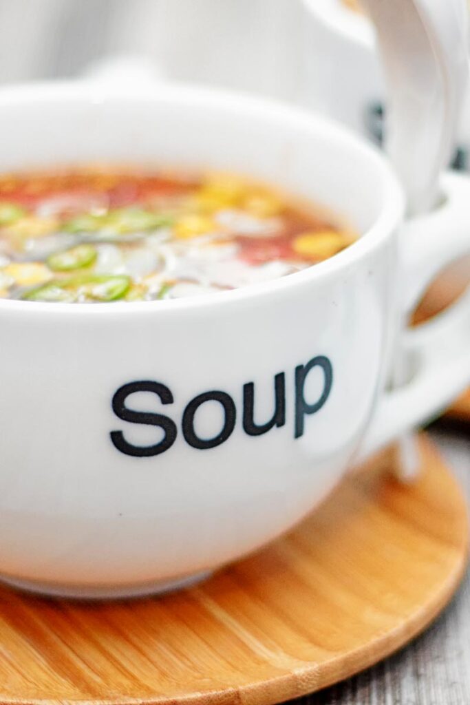 Portrait close up image of a tomato and sweetcorn broth based soup served in a white porcelain cup with soup printed on the front