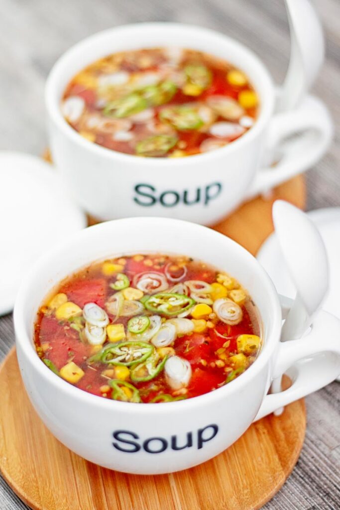 Portrait image of a tomato and sweetcorn broth based soup served in two white porcelain cups with soup printed on the front