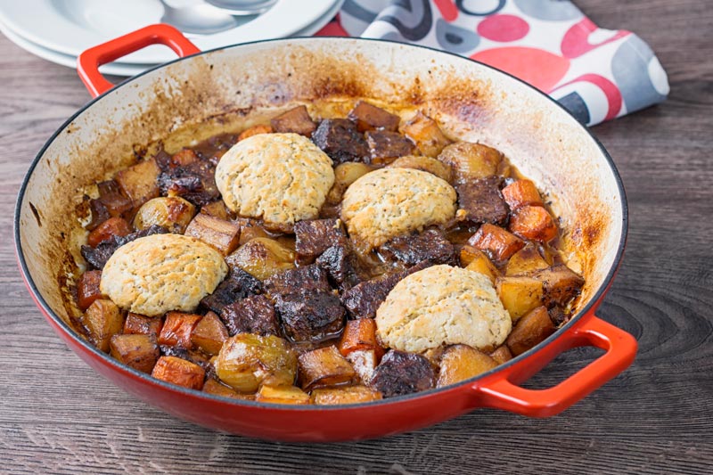 Landscape image of a beef stew with suet dumpling and lots of root vegetables cooked in a shallow cast iron pan