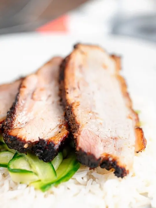 Portrait close up image of sliced crispy roast Chinese pork belly served with rice and pickled cucumbers served on a white plate