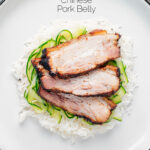 Portrait overhead image of sliced Chinese pork belly served with rice and pickled cucumbers served on a white plate with text overlay
