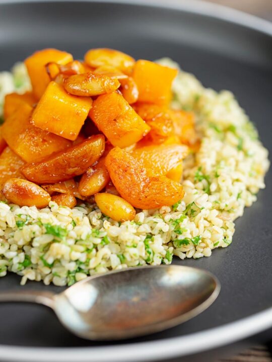 Portrait image of a butternut squash tagine with dried apricots and almonds served on a bed of herbed bulgur wheat on a black plate