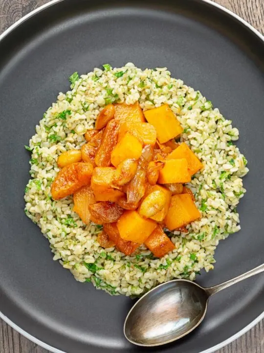 Portrait overhead image of a butternut squash tagine with dried apricots and almonds served on a bed of herbed bulgur wheat on a black plate