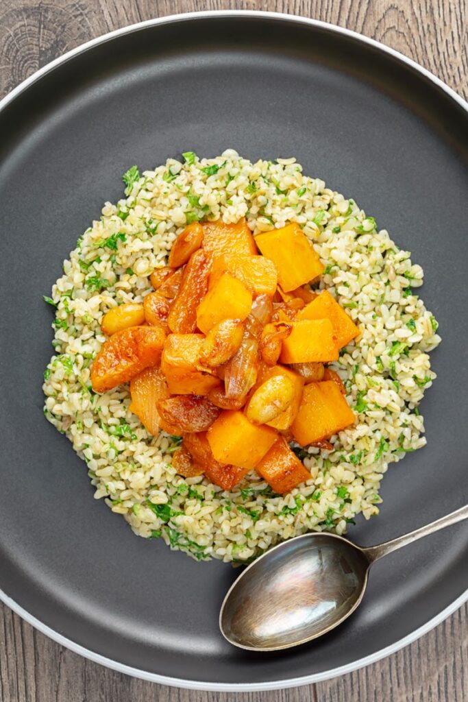 portræt overhead billede af en butternut græskar tagine med tørrede abrikoser og mandler serveret på en seng af herbed bulgur hvede på en sort plade