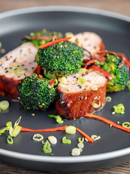 Portrait image of Chinese Char Siu pork tenderloin sliced and served on a dark plate with chilli spring onion and stir fried broccoli