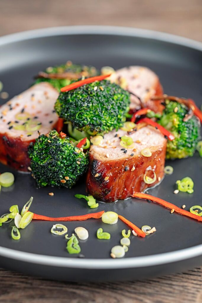 Portrait image of Chinese Char Siu pork tenderloin sliced and served on a dark plate with chilli spring onion and stir fried broccoli