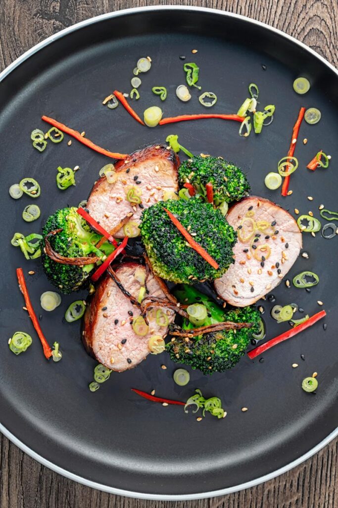 Portrait overhead image of Chinese Char Siu pork tenderloin sliced and served on a dark plate with chilli spring onion and stir fried broccoli