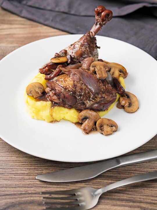 Portrait image of chicken thigh and drumstick in a red wine sauce with mushrooms and bacon served on mashed potato on a white plate