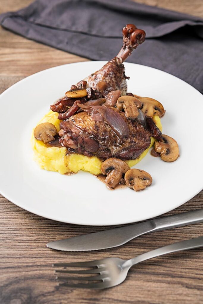 Portrait image of chicken thigh and drumstick in a red wine sauce with mushrooms and bacon served on mashed potato on a white plate
