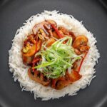 Portrait overhead image of a Chinese chicken and mushroom stir fry served ona black plate with shredded spring onion and white rice