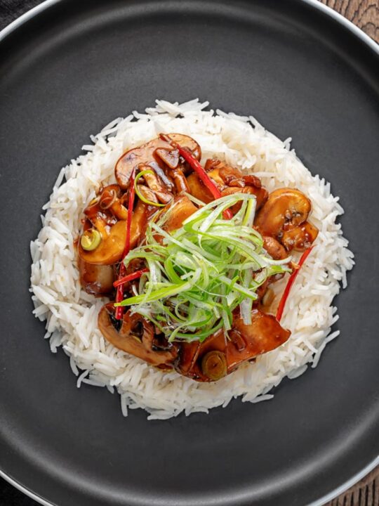 Portrait overhead image of a Chinese chicken and mushroom stir fry served ona black plate with shredded spring onion and white rice