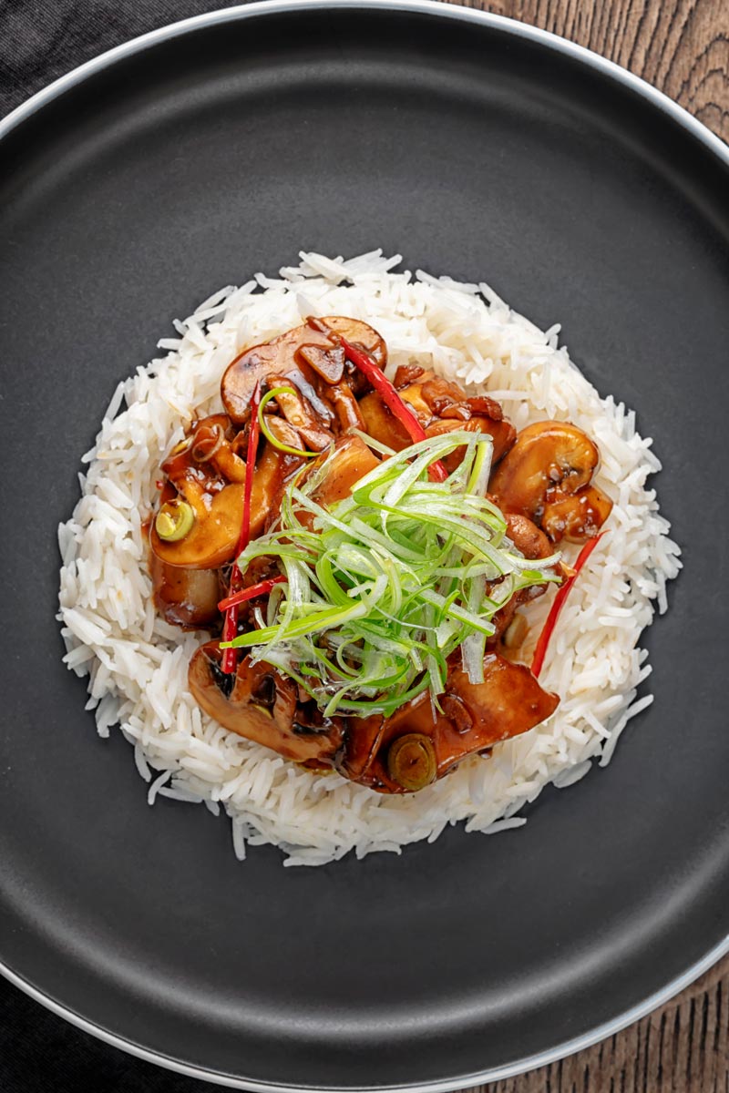 Portrait overhead image of a Chinese chicken and mushroom stir fry served ona black plate with shredded spring onion and white rice